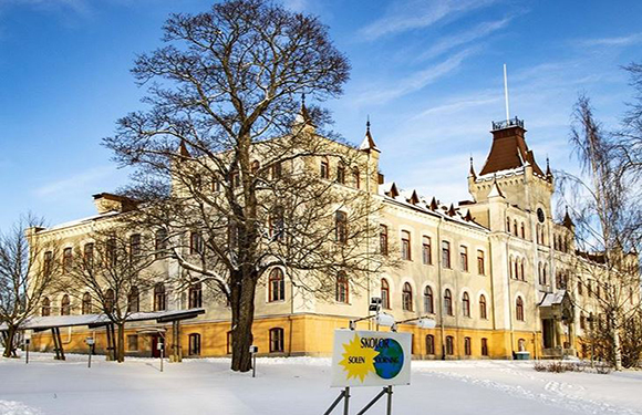 An ornate historical building with a tower and many windows, set in a snowy landscape under a clear blue sky, with a green sign in front.