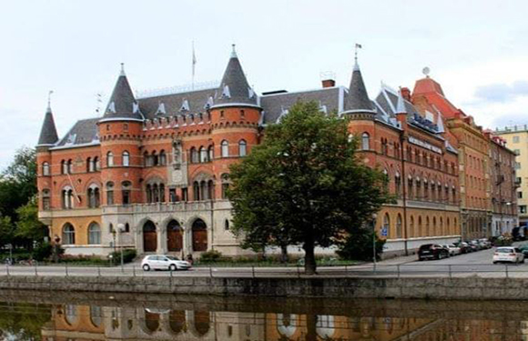 A scenic image of a European-style building with spires by a reflective water body, with trees, clouds, and a visible car.