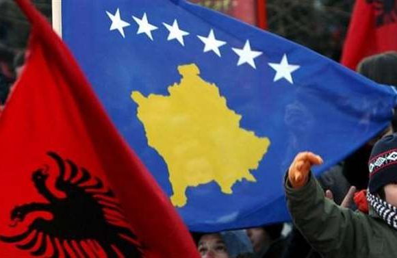 A Kosovo flag with a yellow map and six stars is waved, with a partial view of an Albanian flag and indistinct people behind.