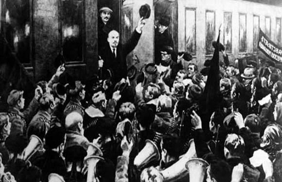 Historical B&W photo of a man addressing a crowd with raised hands and background banners.