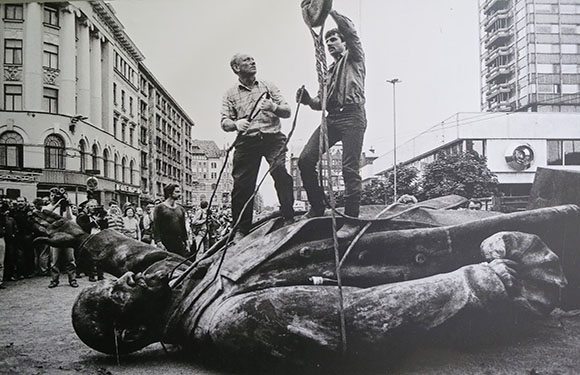 A black and white photo showing two people standing on a toppled statue in an urban setting with onlookers in the background.