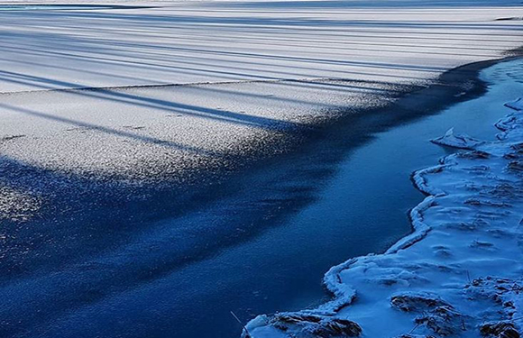 A photo depicts a curved shoreline with a pattern of light and shadows on a frozen lake or river with visible snow and ice.