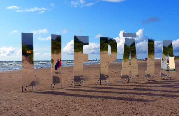 A person standing next to large, reflective, three-dimensional letters on a sandy beach with the sky and trees in the background.