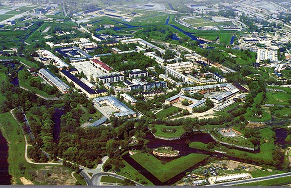 Aerial view shows a large prison complex with various buildings, surrounded by greenery, water bodies, and nearby roads.