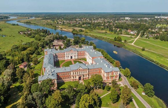 Aerial view of a historic red-brick building with a courtyard, next to a river, surrounded by a park and trees.