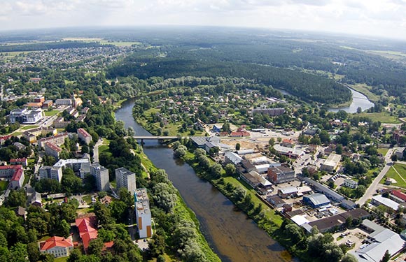 Aerial view of a town with buildings and roads alongside a winding river, surrounded by greenery and trees.