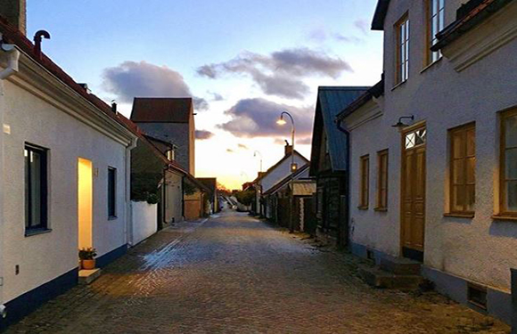 A cobblestone street at dusk with traditional houses on either side and a sunset in the background.