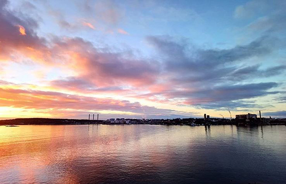 A sunset with vibrant orange and pink hues reflecting on a calm body of water, with silhouettes of distant land and structures on the horizon.