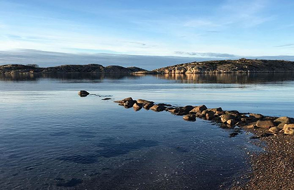 A peaceful landscape with a calm water body, a rock path leading into it, and hills under a clear sky.