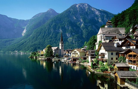 A picturesque lakeside village with traditional houses, a prominent church spire, and lush green mountains in the background.