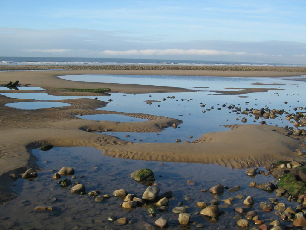 A coastal landscape with tidal pools, sand, and scattered rocks under a partly cloudy sky.