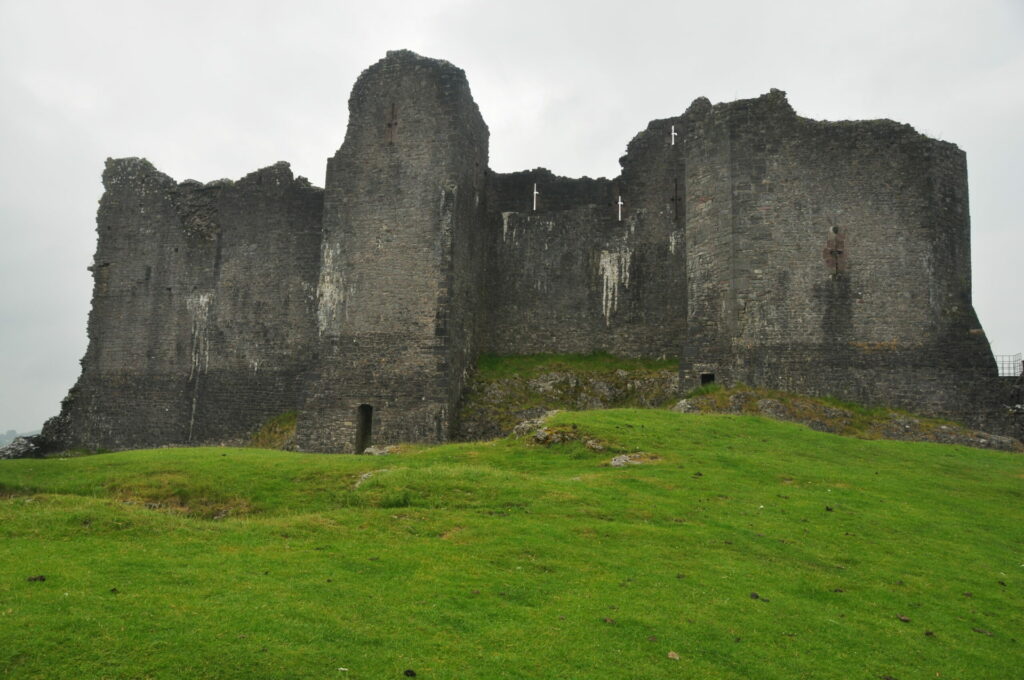 A photo of a large, weathered stone castle with multiple towers and sections of walls standing against a cloudy sky, situated on a grassy field.