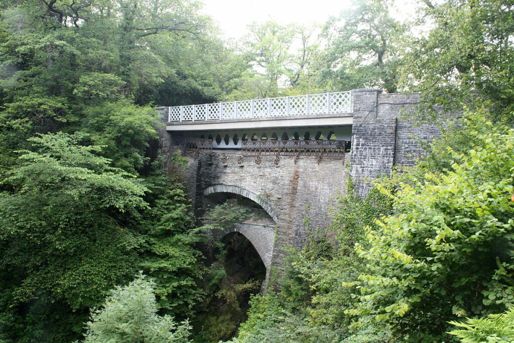 An old stone bridge with a decorative railing, surrounded by lush greenery and trees.