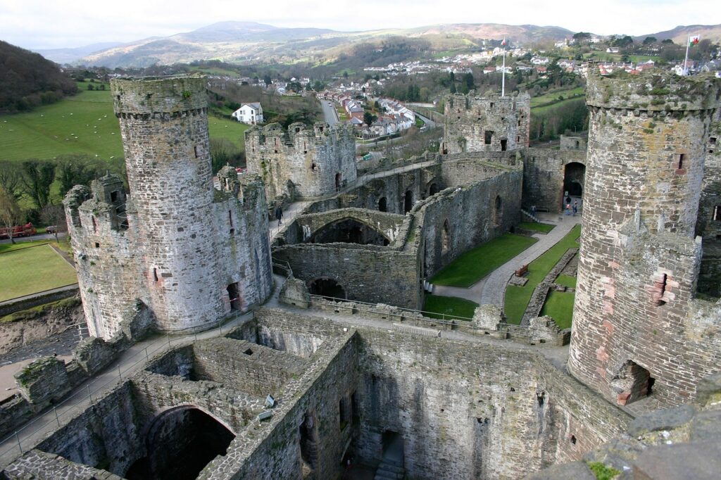 Aerial shot of a ruined medieval castle with stone towers and walls, set against hills and a lawn.