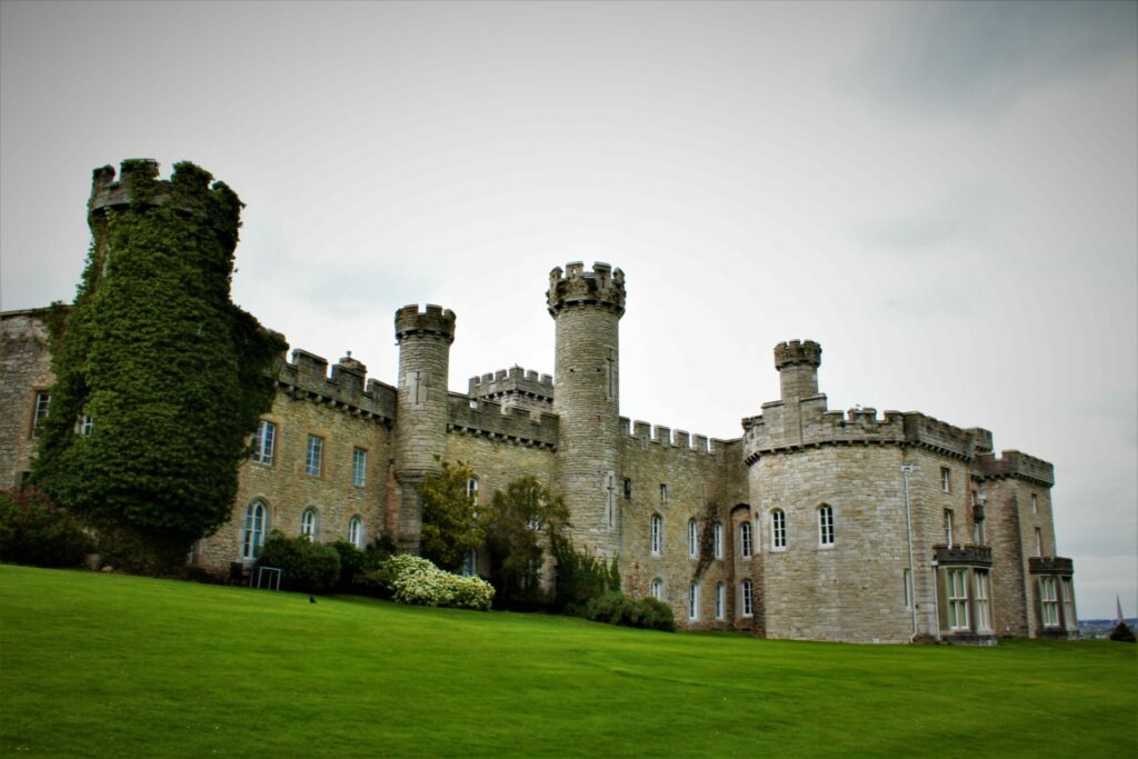 An image of a castle with multiple turrets and a large green lawn in the foreground under an overcast sky.