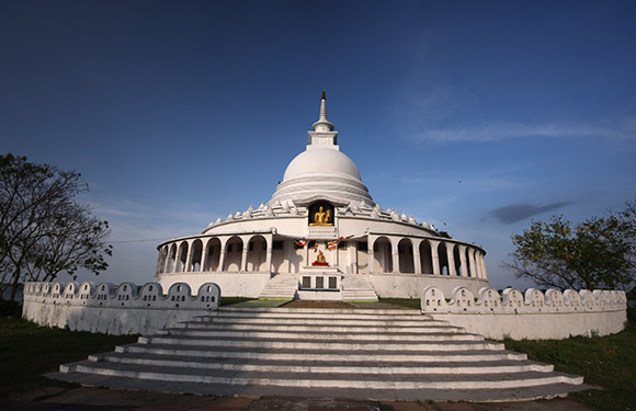 A white domed stupa is set against a lightly clouded blue sky, with trees flanking its series of steps.