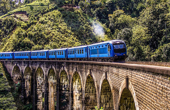 A blue train crossing an arched stone bridge in a lush green landscape with trees and a hill in the background.