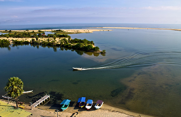 Aerial view of a calm river with a boat creating ripples on the water, adjacent sandy shores with trees, and colorful boats lined up on the bank.