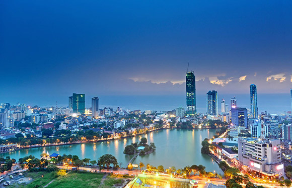 A panoramic view of a city skyline at dusk with illuminated buildings and a river reflecting city lights.