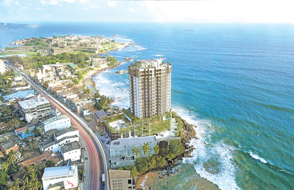 Aerial view of a coastal city with a tall building on a sea cliff, a coastal road, and various smaller buildings.