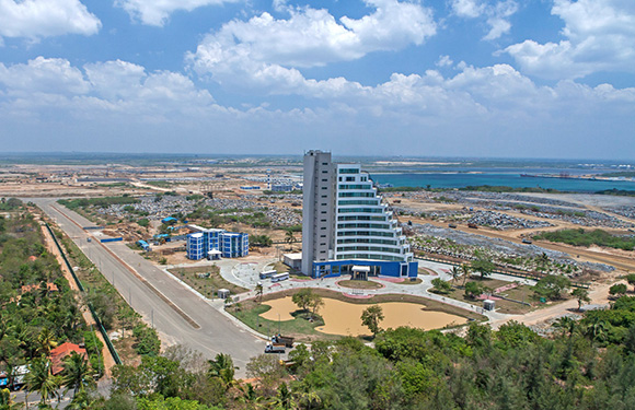 Aerial view of a coastal urban area with a prominent multi-story building, roads, sparse vegetation, and a view of the sea in the distance.