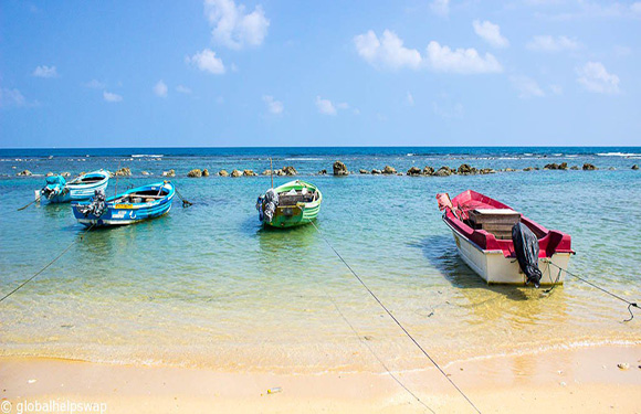 A scenic view of multiple colorful boats floating near the shore of a clear blue sea under a bright blue sky with sparse clouds.