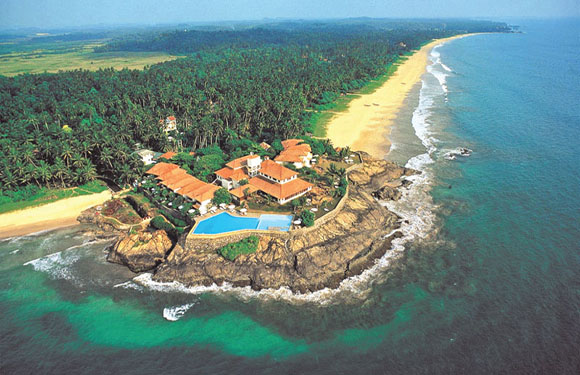 Aerial view of a coastal resort with a swimming pool, surrounded by a lush green landscape and bordered by a sandy beach and the blue ocean.