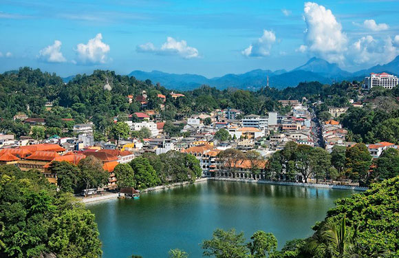 A scenic view of a town with traditional buildings beside a calm lake, surrounded by lush greenery and hills under a blue sky with some clouds.