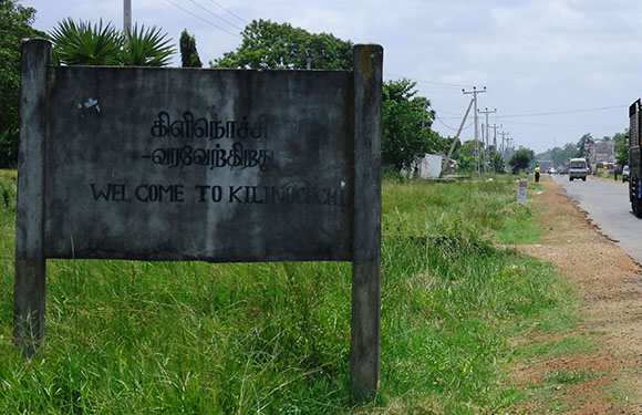 A weathered bilingual Tamil-English welcome sign stands roadside, with a grassy backdrop and a long road ahead.