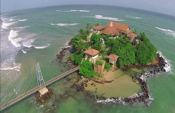 Aerial view of a small island with buildings surrounded by a sea, connected to the mainland by a narrow pier.