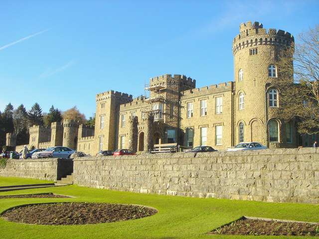 A photo of a castle with a cylindrical tower, surrounded by a well-manicured lawn and parked cars under a clear blue sky.