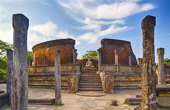 A photograph of ancient ruins with stone pillars and a circular structure in the center under a blue sky with clouds.