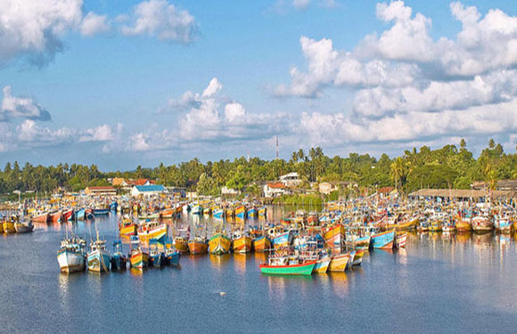 A colorful array of boats densely packed in a calm waterway, with greenery and a blue sky with scattered clouds in the background.