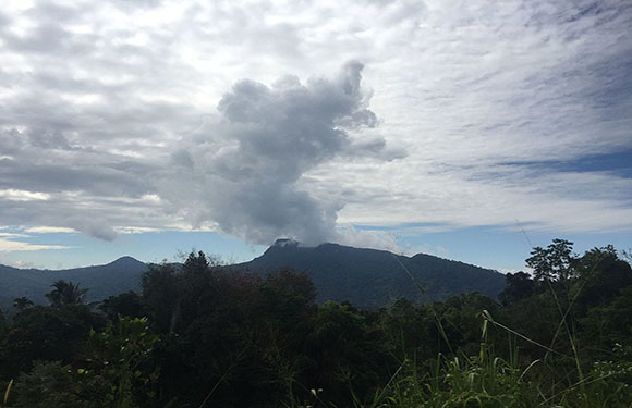 A landscape image shows a mountain range with smoke indicating volcanic activity, under a partly cloudy sky with greenery in front.