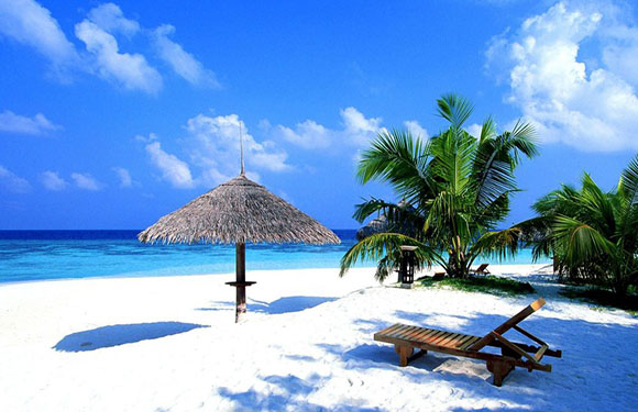 A tropical beach scene with white sand, a single wooden sun lounger, a thatched umbrella, clear blue sky, and turquoise water.