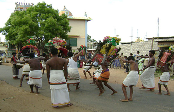 People in traditional attire perform a street dance, some with colorful headgear, against a backdrop of buildings and trees.