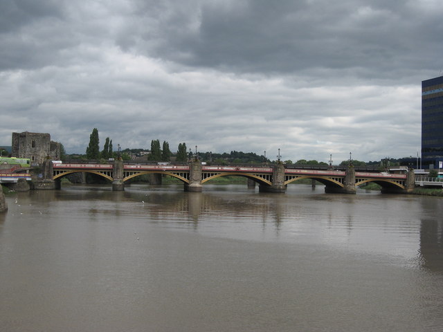 A bridge with multiple arches spans over a wide river under a cloudy sky. Buildings are visible on the right, and trees line the far bank.