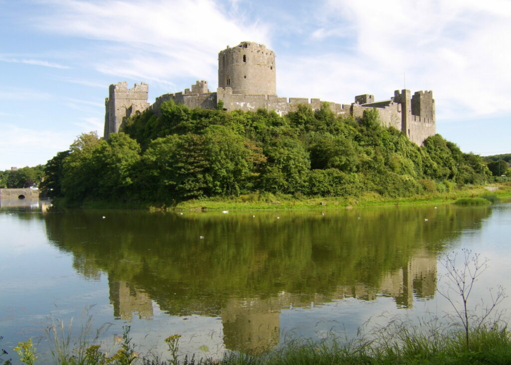 A medieval castle with round towers sits by a calm river, its reflection visible in the water, amidst greenery under a clear sky.