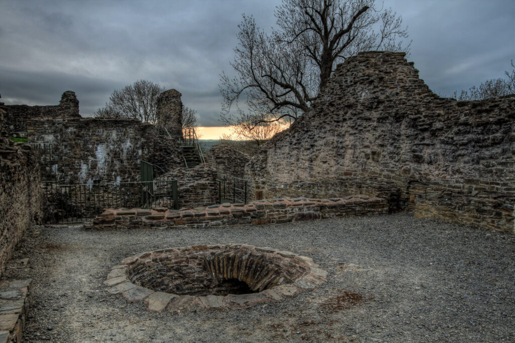 A dusk image of ancient ruins with a circular pit, crumbling walls, and a bare tree against a cloudy sky.