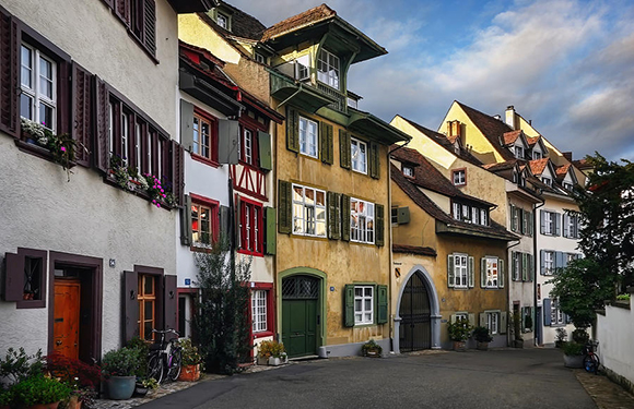 A picturesque street with traditional European buildings featuring colorful facades, shuttered windows, and flower boxes under a clear sky.