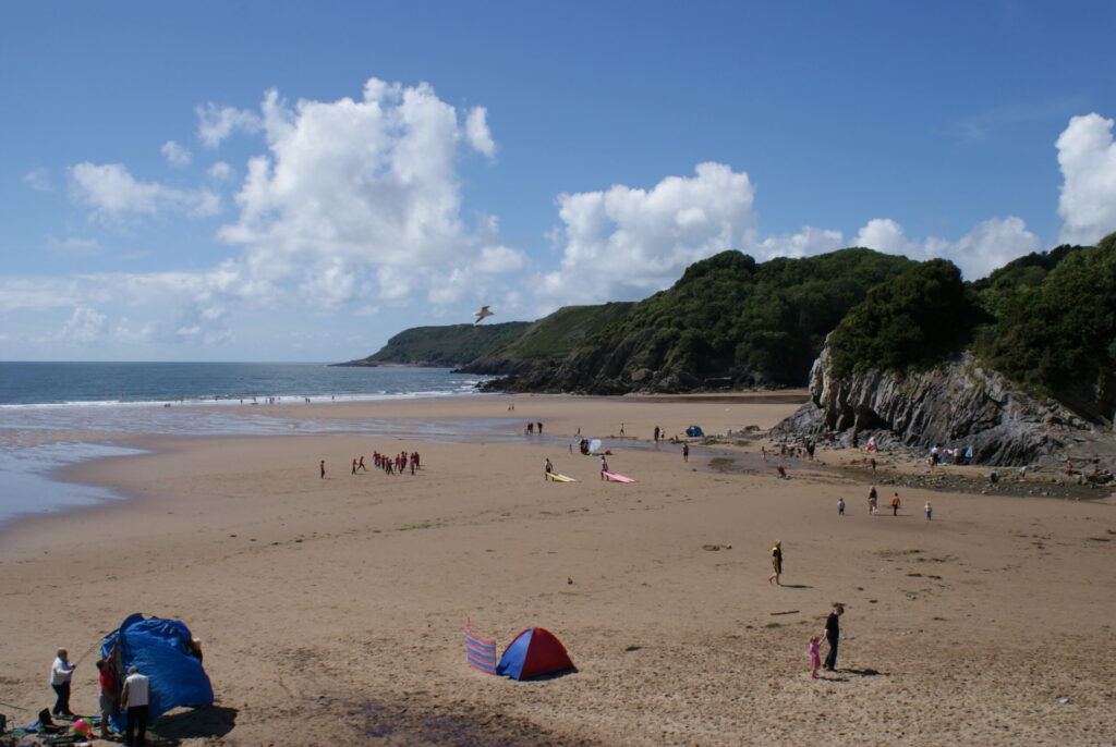 A sandy beach with people, colorful tents, a tree-covered headland to the right, and a clear, cloudy sky above.