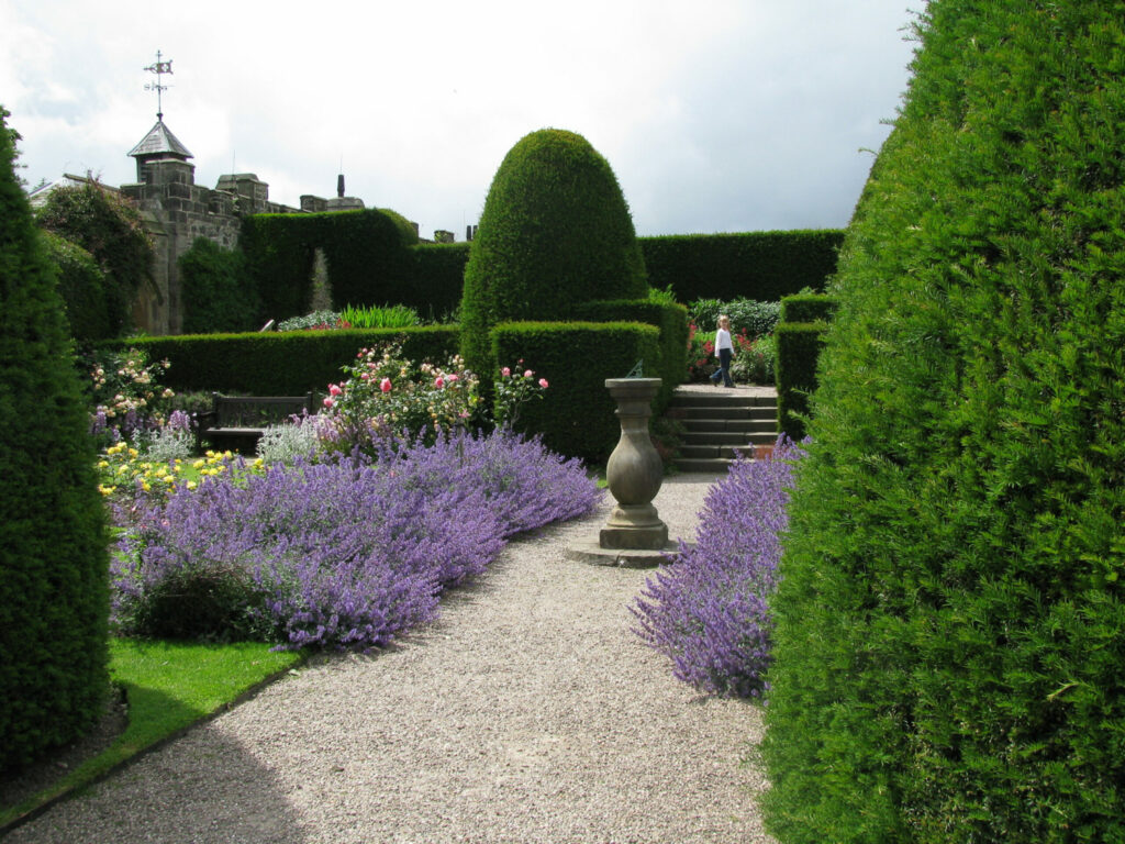 A garden pathway lined with purple flowers and neatly trimmed green hedges, with a decorative urn on a pedestal to the left and a cloudy sky above.