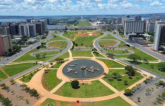 Aerial view of a city park with a central fountain, surrounded by paths, greenery, and buildings under a cloudy blue sky.