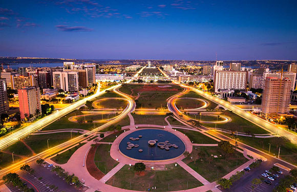 Aerial dusk view of a city with lit streets, a water fountain, buildings on both sides of an open space leading to the horizon.