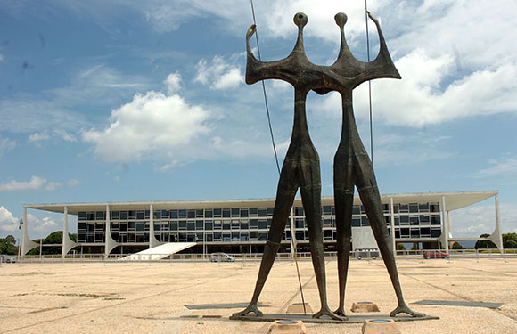 A sculpture of four abstract, crowned figures stands before a modernist building with a flat facade under a cloudy sky.