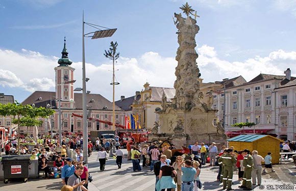 A bustling town square with a baroque monument, European buildings, and a clear sky backdrop.