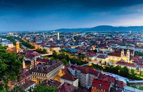 Aerial view of a city at dusk with illuminated buildings and streets, with mountains in the background under a cloudy sky.