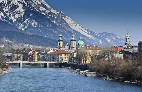 A scenic view of a city with historical buildings and a bridge over a river, with snow-capped mountains in the background.