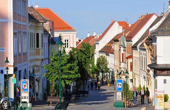 A scenic street with vibrant European buildings, pedestrians, and interspersed green trees under a clear sky.