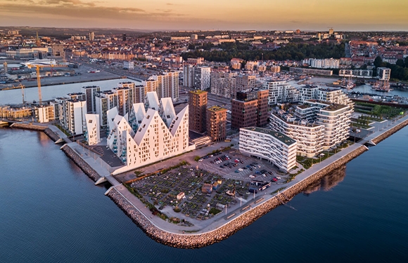 Aerial view of a modern waterfront urban area during sunset with distinct architectural buildings and a clear sky.